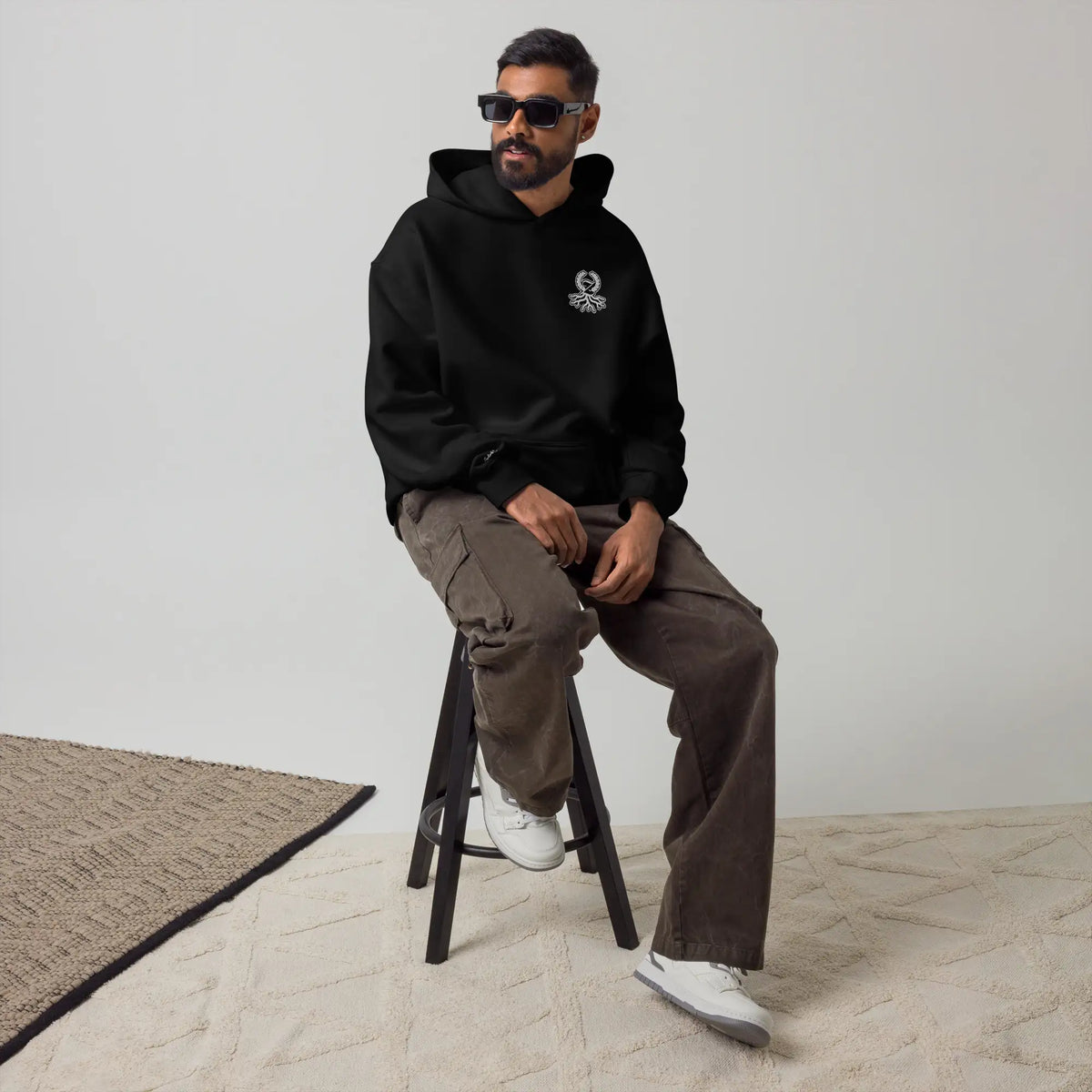 Hombre con sudadera oversize negra Zaylachi Co. 319gsm sentado en taburete. Streetwear urbano premium unisex.