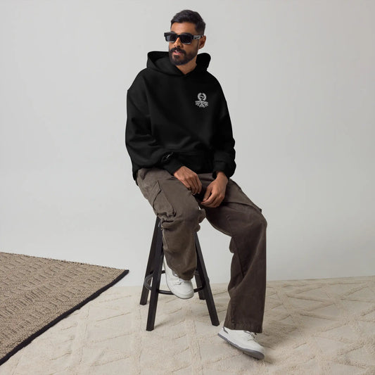 Hombre con sudadera oversize negra Zaylachi Co. 319gsm sentado en taburete. Streetwear urbano premium unisex.